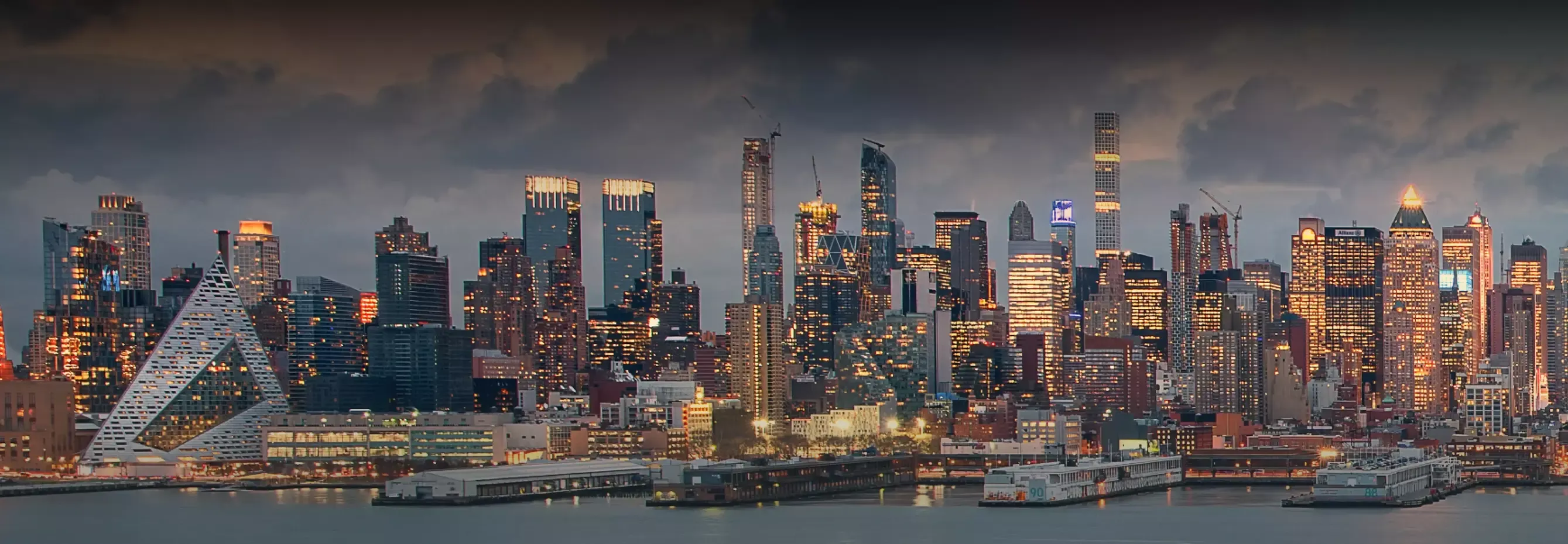 Twilight cityscape with illuminated buildings and a the distinct pyramid-shaped VIA 57 WEST apartment building by the waterfront.