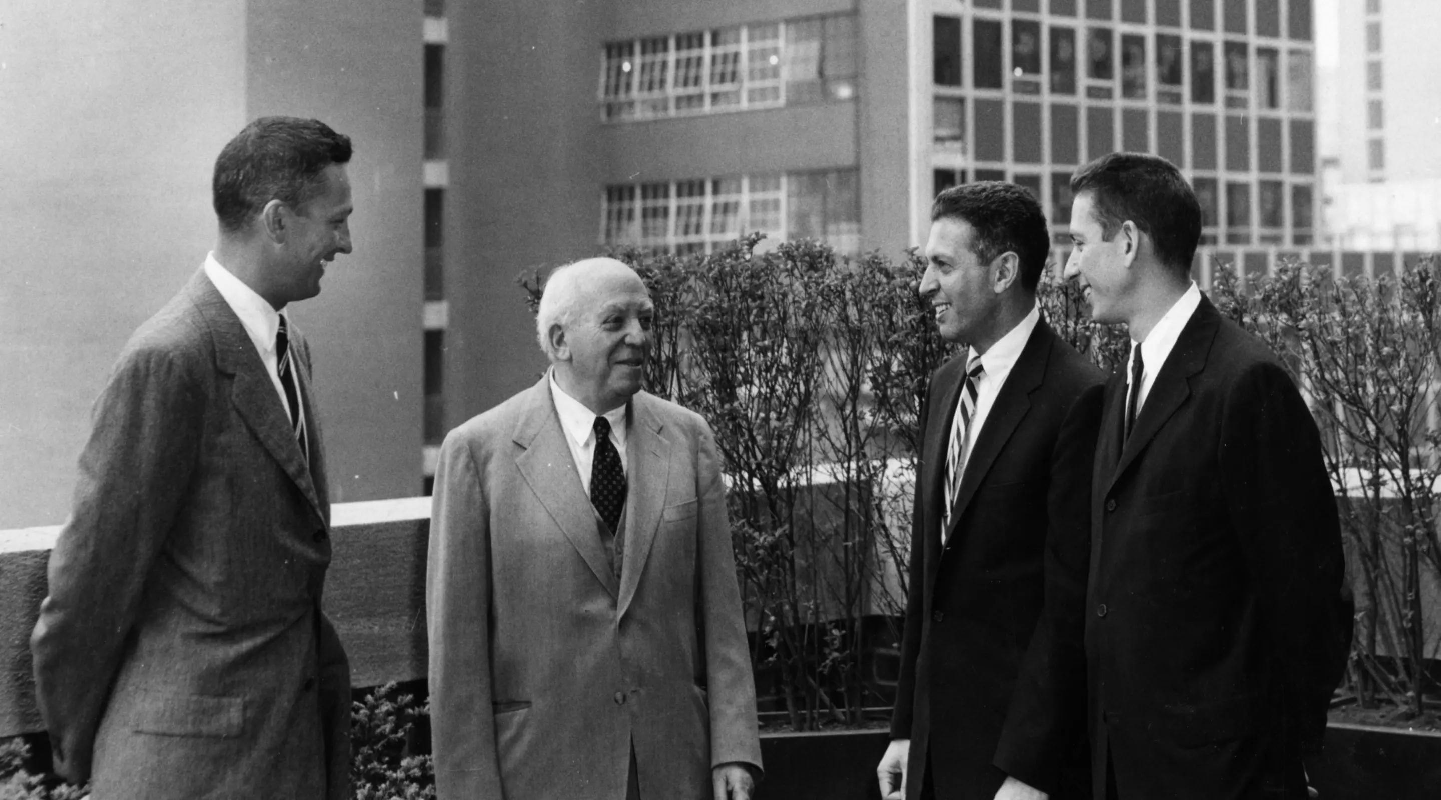 Black and white photo of four men conversing on a rooftop, city buildings in the background.