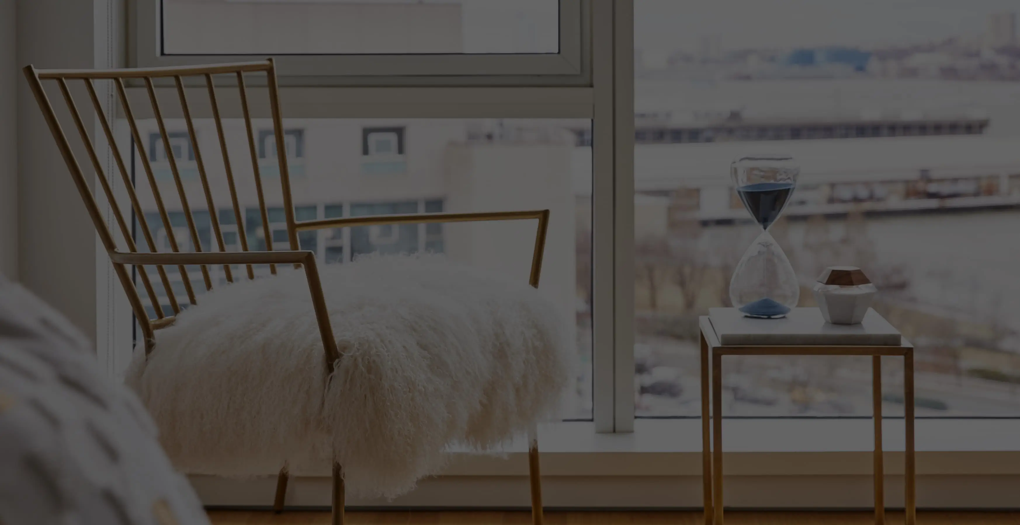 Cozy nook with a furry throw on a gold chair, hourglass on side table, and a riverside view.